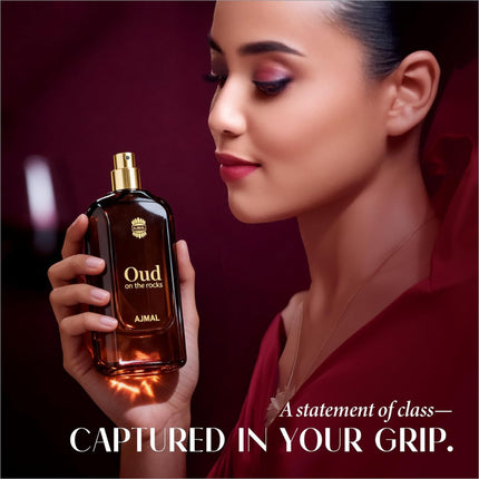 A dark amber glass bottle with a gold-tone spray nozzle of ajmal oud on the rocks eau featuring smoky oud and woody amber notes